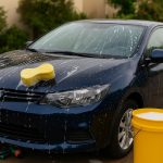 Person washing a car in a driveway with soap, sponge, and water for home car cleaning tips
